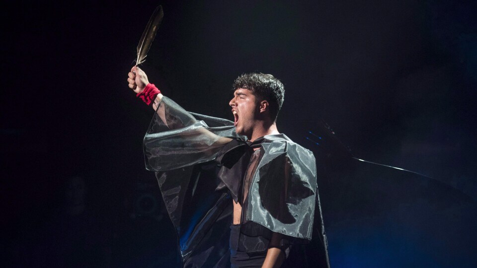 De profil, un homme chante. Il tient à bout de bras une longue plume. Il porte une cape faite d'un tissu transparent.