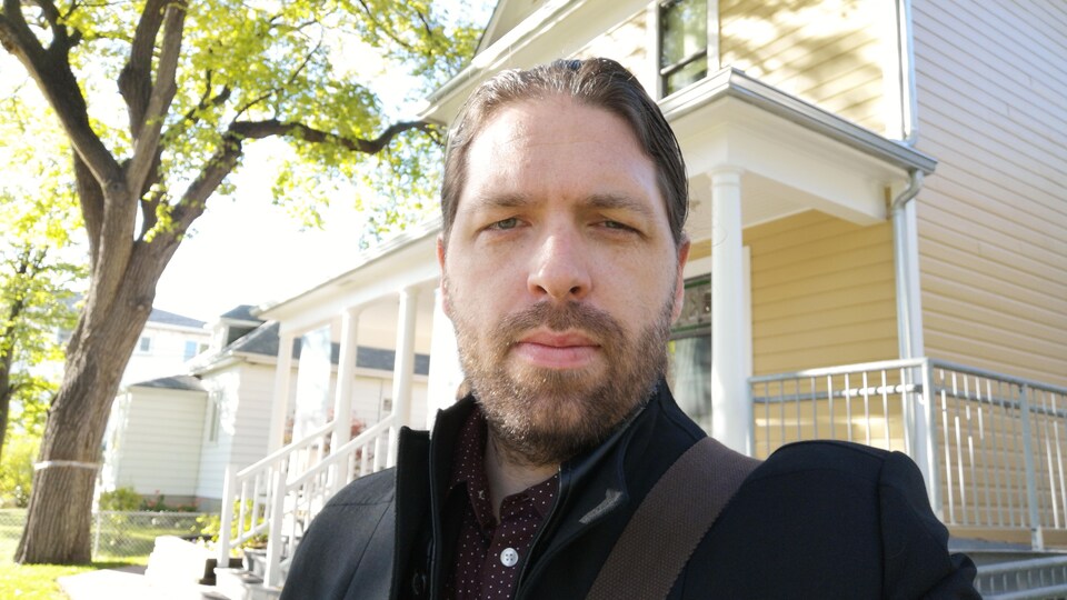 Portrait de Jérôme Melançon. Il est dehors devant une maison. Il y a un arbre à sa gauche.