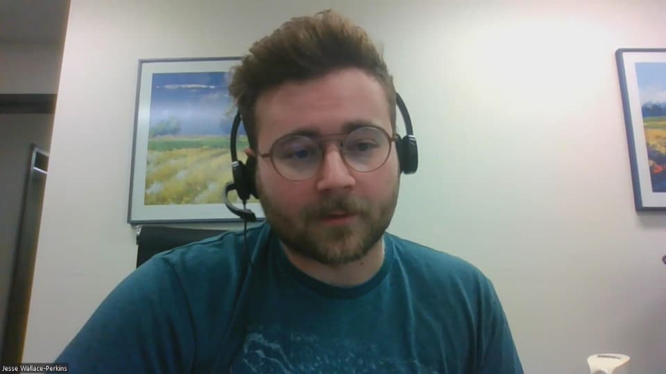 Un homme avec une barbe et des lunettes porte un casque d'écoute. Il répond aux questions de la journaliste par visioconférence.