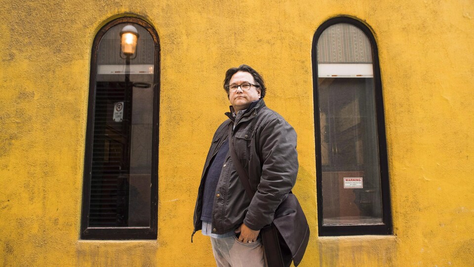 Un homme portant des lunettes et une veste noire pose devant un bâtiment.