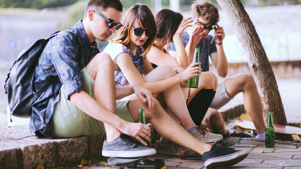 Deux jeunes hommes et deux jeunes femmes qui semblent d'âge mineur sont assis sur une chaîne de trottoir avec des skateboards et des bouteilles de bière.