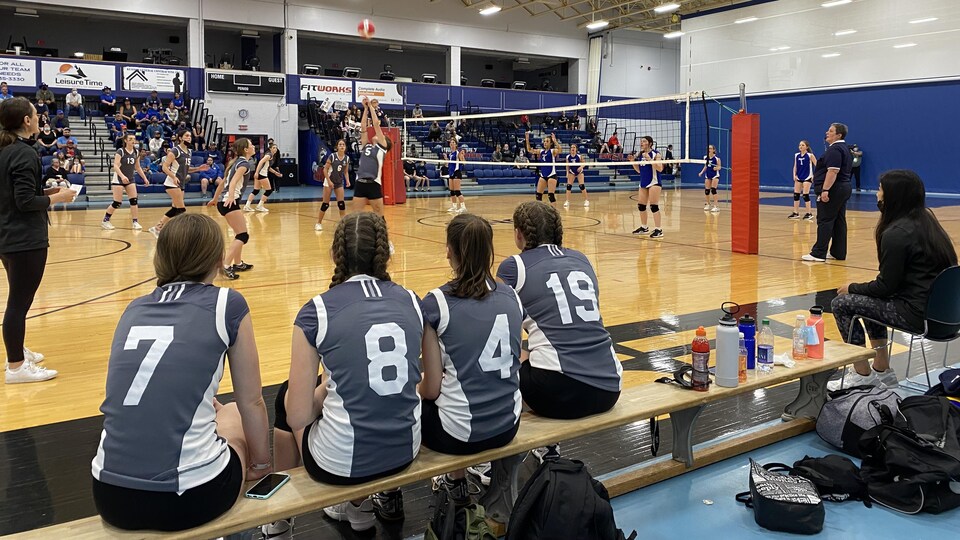 Des joueuses de volleyball portant des maillots gris sont assises sur un banc en bois et regardent leurs coéquipières qui disputent un match dans un gymnase.