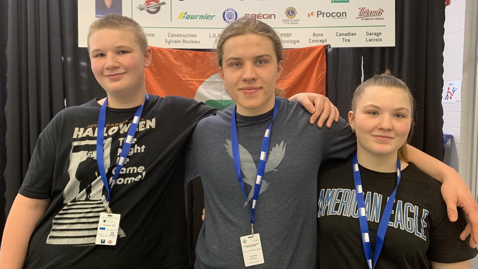 Two brothers and a sister are photographed, arm in arm, in front of the official flag of the Quebec Games.