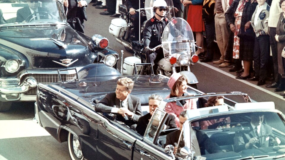 John F. Kennedy est assis à l'arrière d'une en voiture décapotable accompagné de son épouse lors d'un défilé, escorté par un policier à moto.