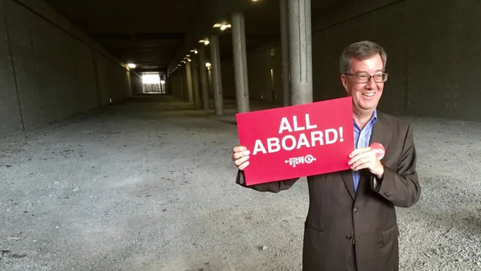 Jim Watson, debout au début d'un tunnel, tenant dans ses mains une affiche indiquant All Aboard! ou Tous à bord!. 