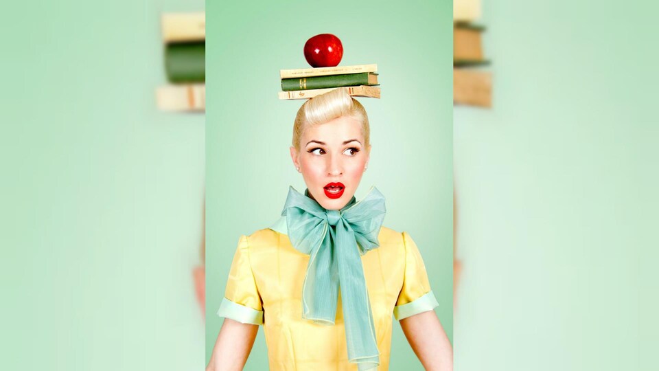 Une femme costumée dans le style pin up tient des livres et une pomme en équilibre sur sa tête.