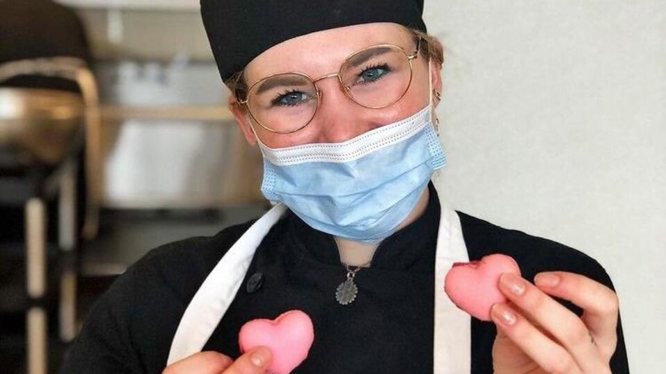 Une jeune femme avec des lunettes et un masque sanitaire tient deux macarons roses en forme de coeur dans ses mains.