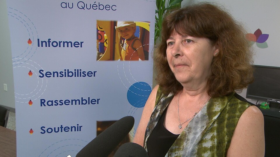 Une femme aux cheveux châtains et aux yeux verts répond aux questions des journalistes devant une affiche publicitaire de la Fédération québécoise de l'autisme.
