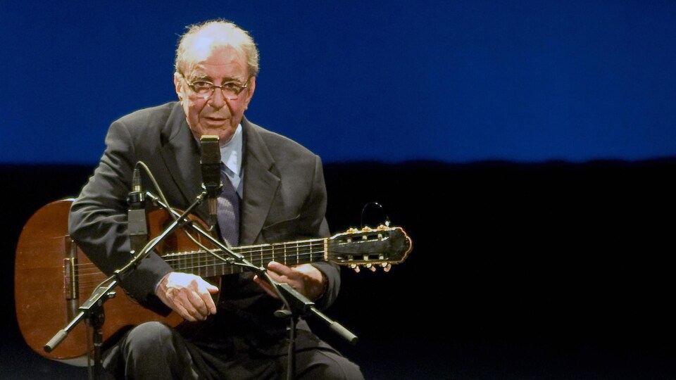 Le chanteur est assis face au micro, une guitare à la main, sur scène.