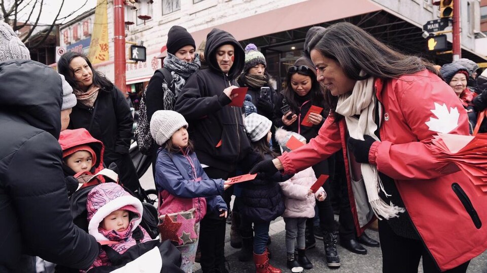 Une femme dans la quarantaine en veste rouge avec une feuille d'érable donne des feuilles rouges à des enfants et des adultes dans une rue lors d'un défilé.