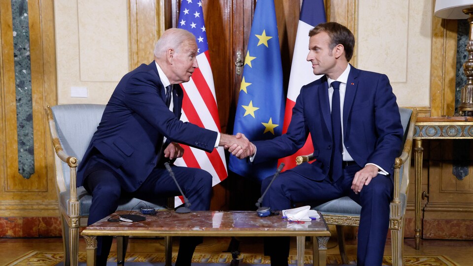 Joe Biden y Emmanuel Macron, ambos sentados, dándose la mano,