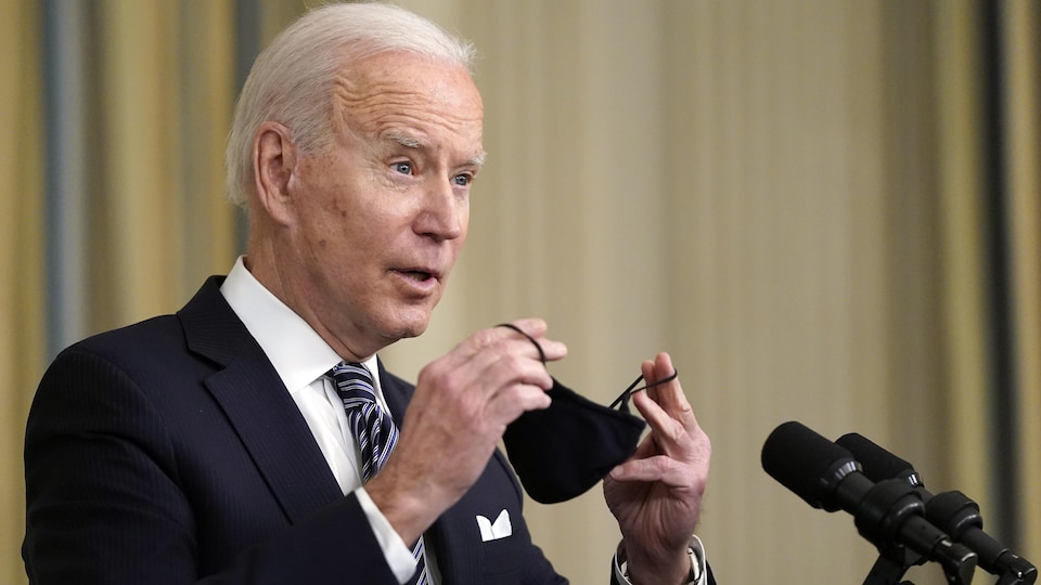 Le président américain, Joe Biden, s'apprête à revêtir un couvre-visage dans une salle, devant des micros.