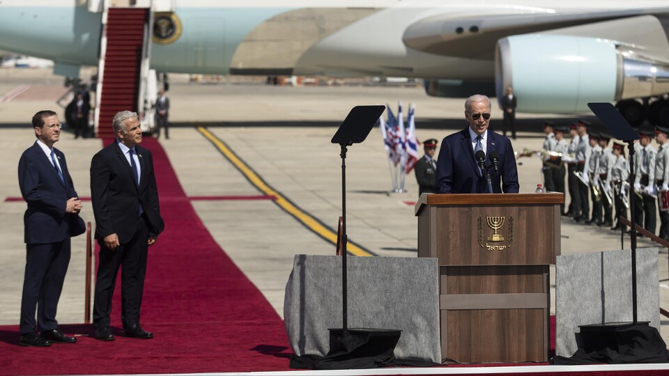 Joe Biden prononce un discours sur le tarmac d'un aéroport. Deux interlocuteurs l'écoutent.