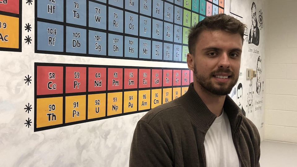 Portrait d'un jeune homme dans une salle de classe. Il est placé devant un tableau périodique de chimie géant
