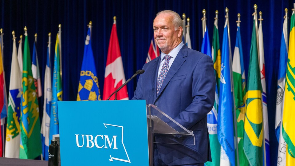 John Horgan parle au micro d'un pupitre devant des drapeaux lors du Congrès de l’Union des municipalités de la C.-B.