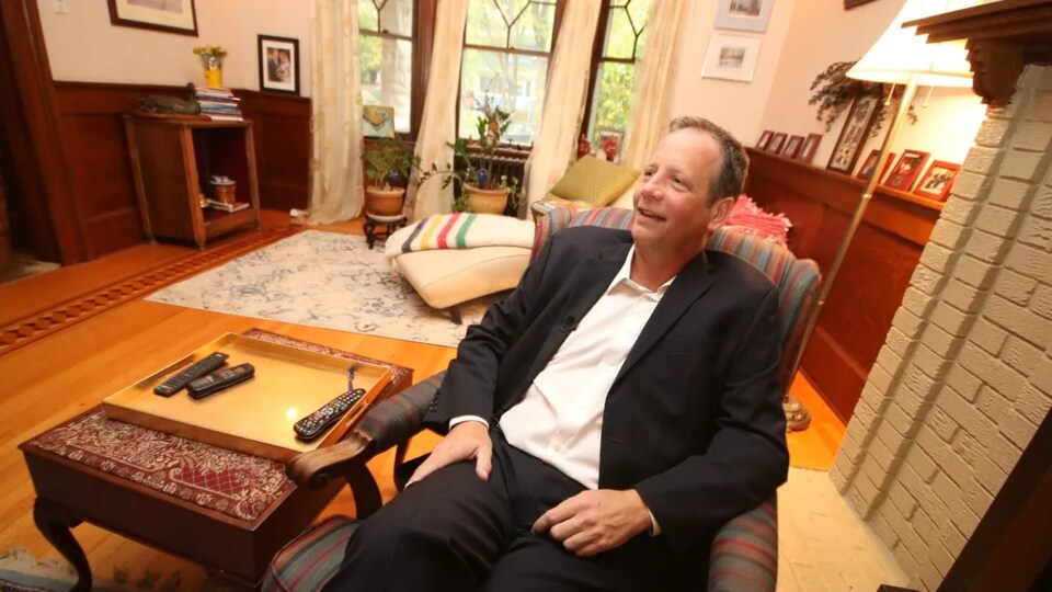 John Orlikow est assis sur un fauteuil dans le salon d'une maison.