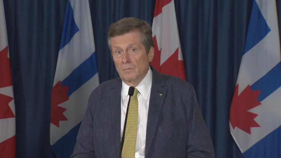 Le maire de Toronto, John Tory, assis devant une table et un micro. Derrière lui, une série de grands drapeaux.