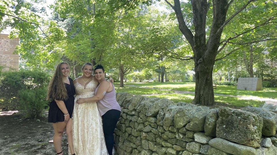 Jolène, en robe de bal de finissante, est accompagnée de sa mère et de sa soeur. Elles prennent une photo dans un parc. 