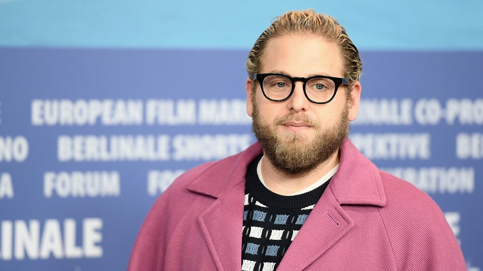 L'homme barbu pose pour les photographes devant un mur. Il sourit et porte des lunettes.