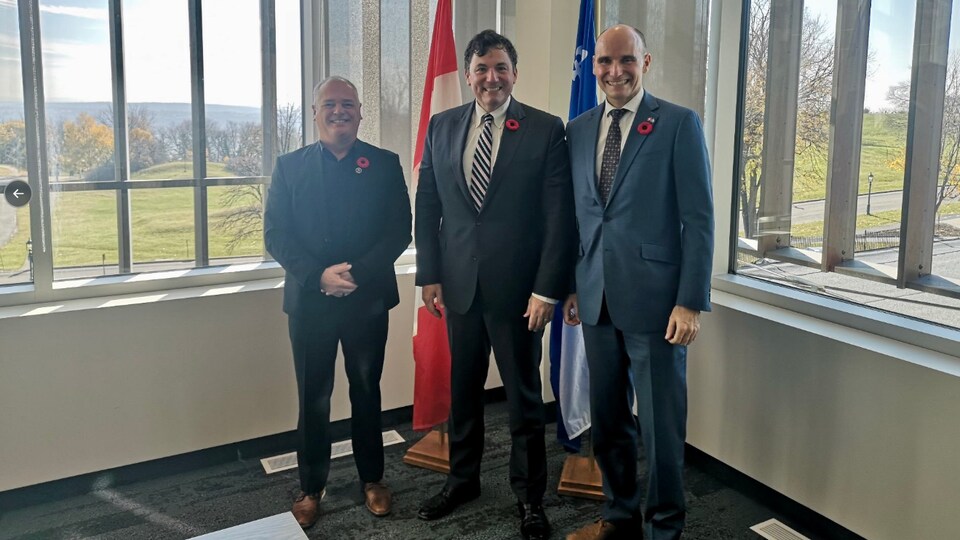 Un plan large de trois hommes souriants l'un à côté de l'autre devant des drapeaux du Québec et du Canada.