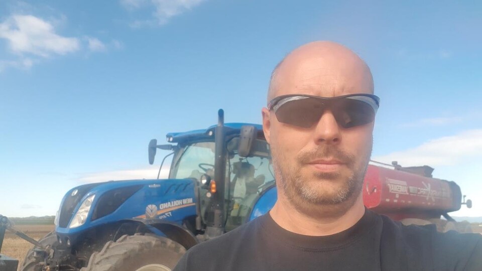 Un homme portant des lunettes fumées dans un champ devant un tracteur bleu.