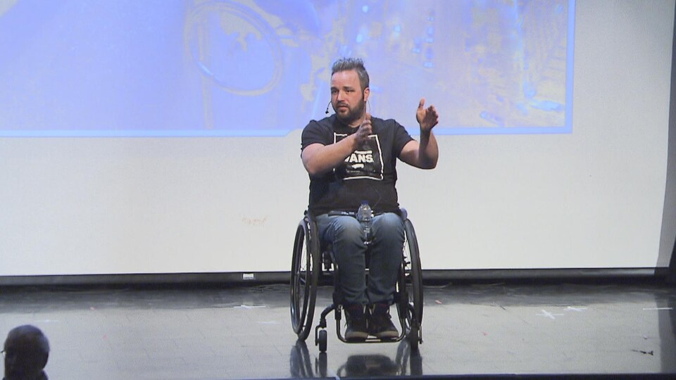Le conférencier en santé et sécurité au travail Jonathan Plante livrant un témoignage à des étudiants, sur la scène, en chaise roulante