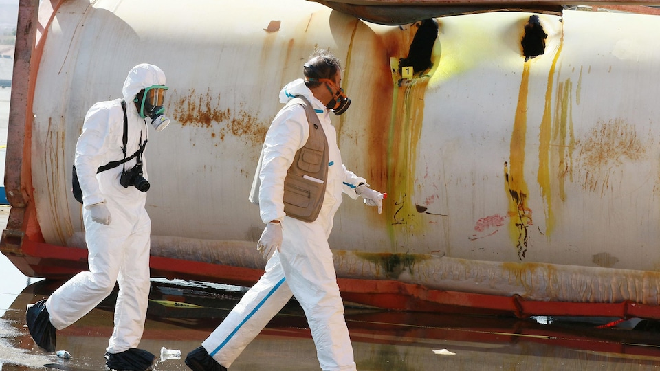 Vêtus de combinaisons intégrales blanches et équipés d'appareils respiratoires, deux experts passent devant le conteneur endommagé.