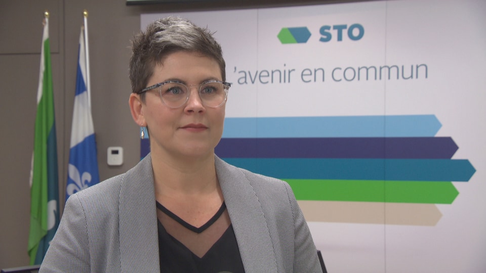 La femme se tient bien droite devant des drapeaux du Québec et de la société des transports ainsi qu'une affiche sur laquelle on peut lire « STO L'avenir en commun ».