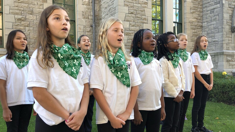 Les jeunes filles chantent debout l'une à côté de l'autre, vêtues d'un chandail blanc ainsi que d'un foulard vert et blanc aux couleurs du drapeau.