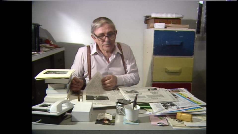 Un homme dans la soixantaine assis à un bureau consulte un journal.