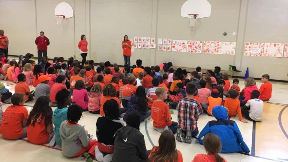 Des élèves portant un chandail orange, pour la plupart, sont assis dans un gymnase et écoutent des adultes, debout, devant eux. (archives)