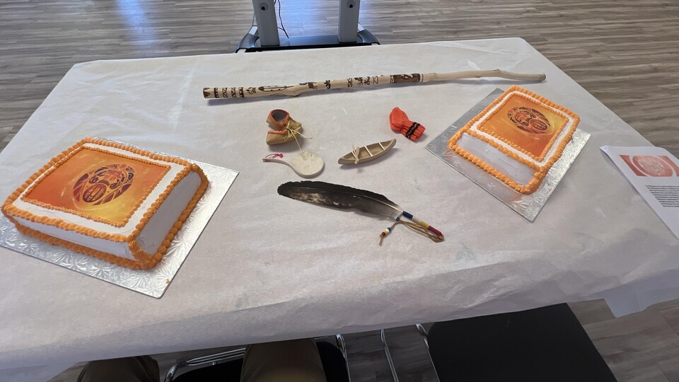 Deux gâteaux au crémage orange sont déposés sur un table. Au milieu, un plume d'oiseau, un canot miniature, une paire de chaussettes orange, une paire de mocassins et un bâton de bois