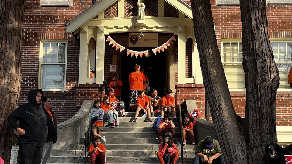 Des jeunes portent des chandails orange et se tiennent debout devant l'entrée de l'école catholique St Thomas Aquinas.  