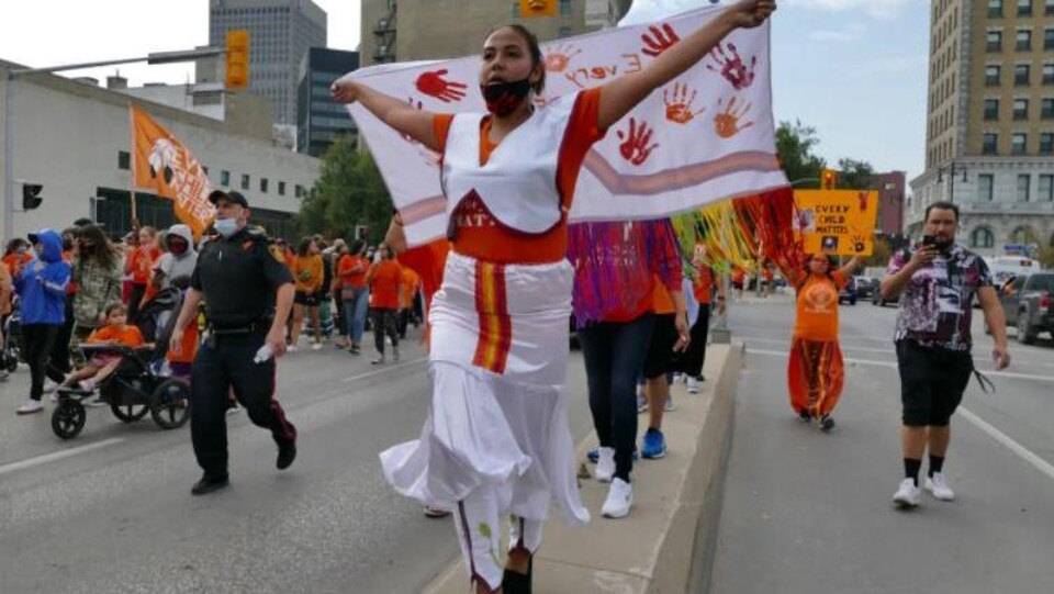 Plusieurs personnes marchent dans les rues de Winnipeg vêtues en orange et portant drapeaux et affiches pour soutenir la réconciliation avec les peuples autochtones.