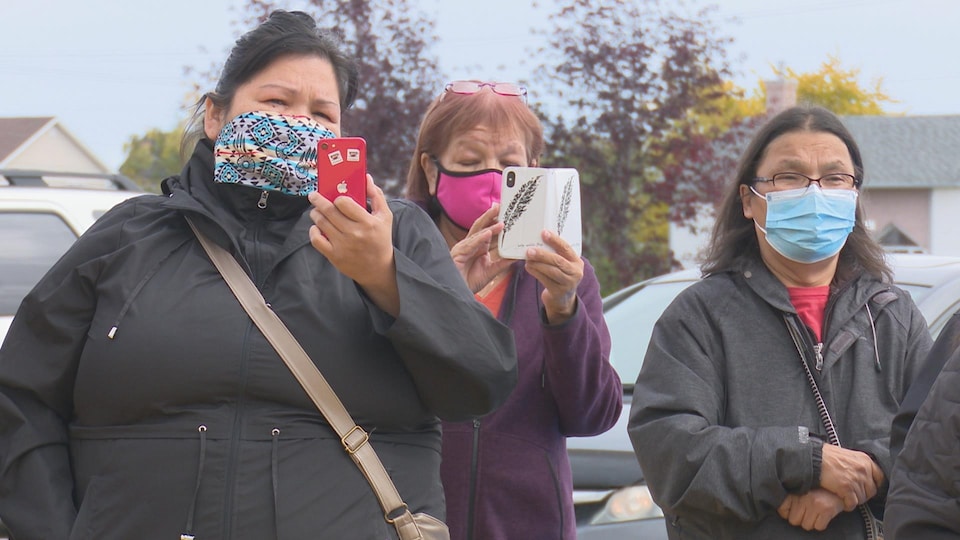 Trois femmes masquées, debout dehors, dont deux regardent leur téléphone cellulaire.