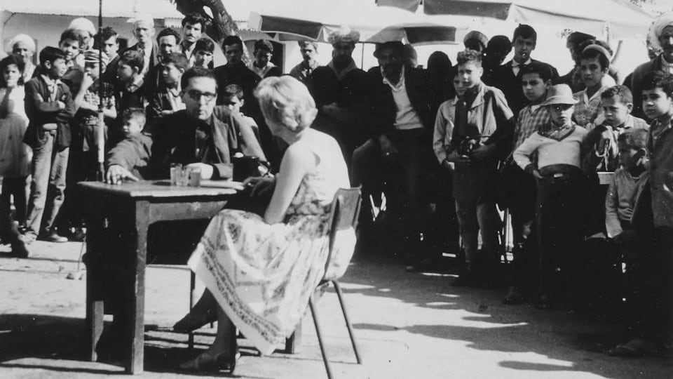 Judith Jasmin, assise à un pupitre entourée d'une foule au milieu d'une place publique en Algérie.