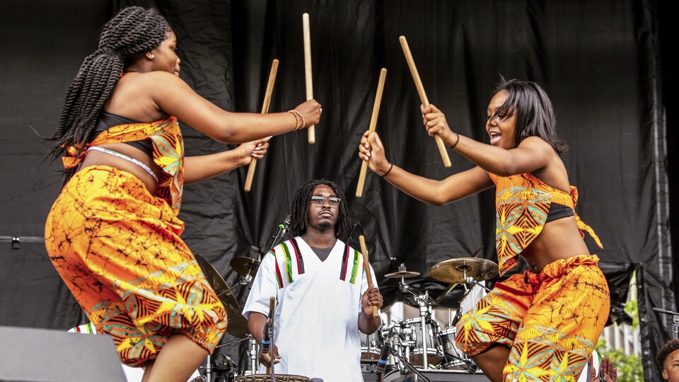 Deux femmes noires dansent avec des bâtons, accompagnées par un joueur de tam-tam.