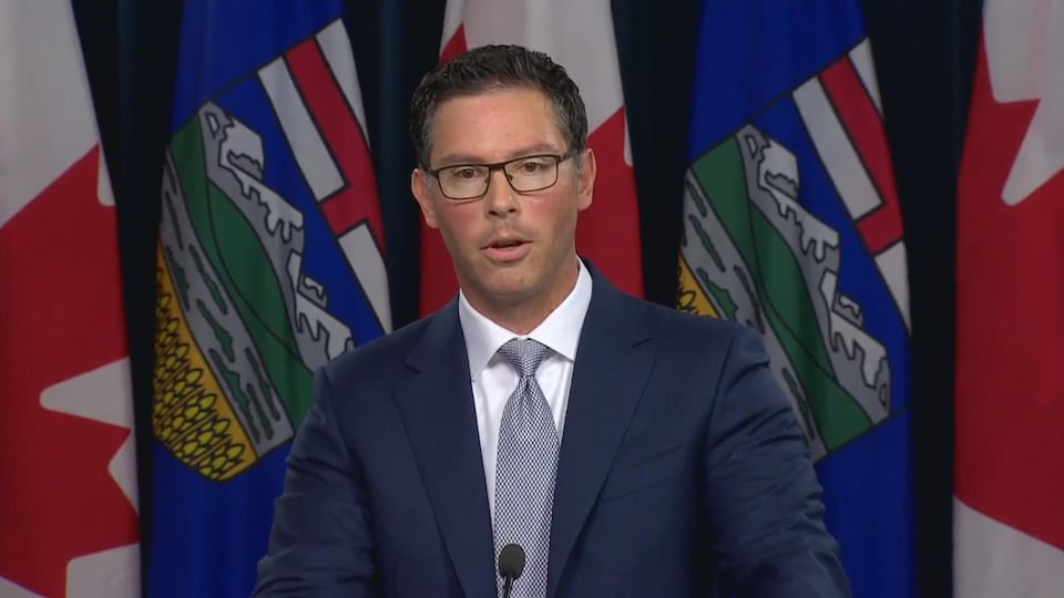 Un homme en costume cravate parle au podium devant une rangée de drapeaux de l'Alberta et du Canada. 