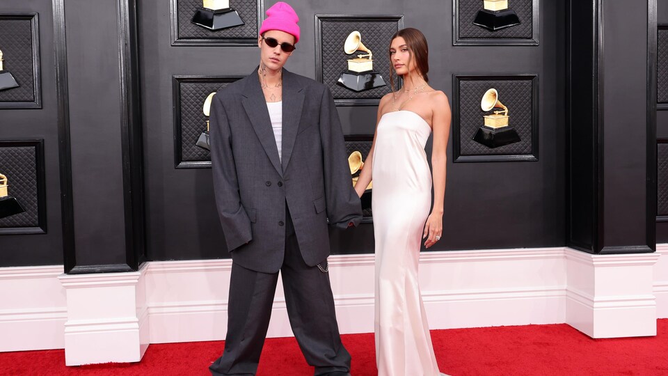 Un homme dans un habit trop grand est accompagné d'une femme habillée en blanc sur le tapis rouge des Grammys. 