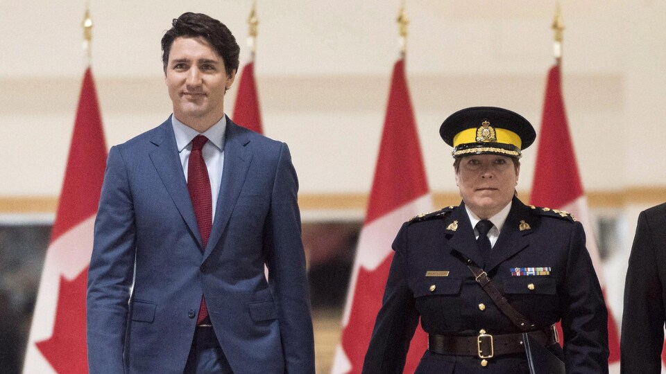 Justin Trudeau en complet et Brenda Lucki en uniforme de la GRC marchent côte à côte en direction du photographe. Des drapeaux du Canada se trouvent derrière eux, au fond de la pièce.