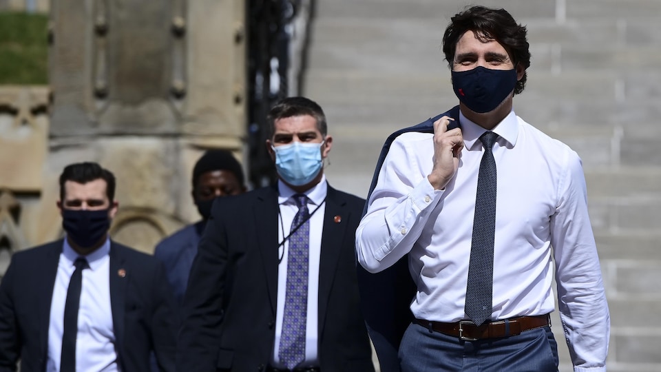 Justin Trudeau, portant un masque, marche dehors tenant son veston sur son épaule et suivi par plusieurs personnes.