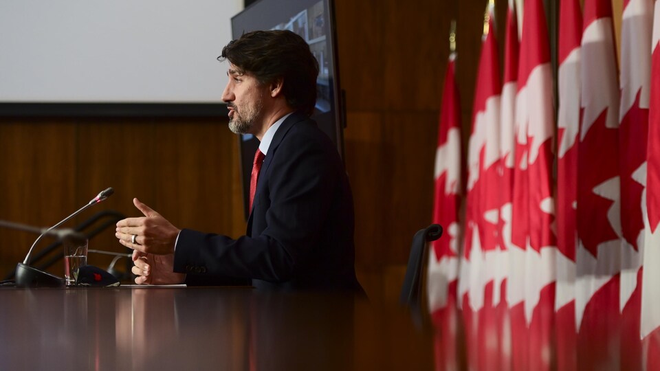 Justin Trudeau de profil et assis à une table avec derrière lui des drapeaux du Canada.