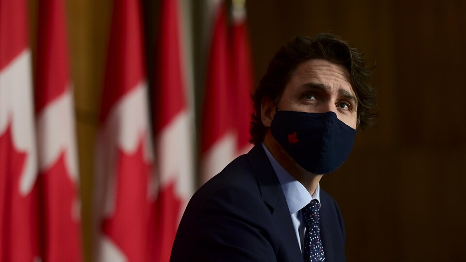 Justin Trudeau, portant un masque, assis devant des drapeaux canadiens pour une conférence de presse.