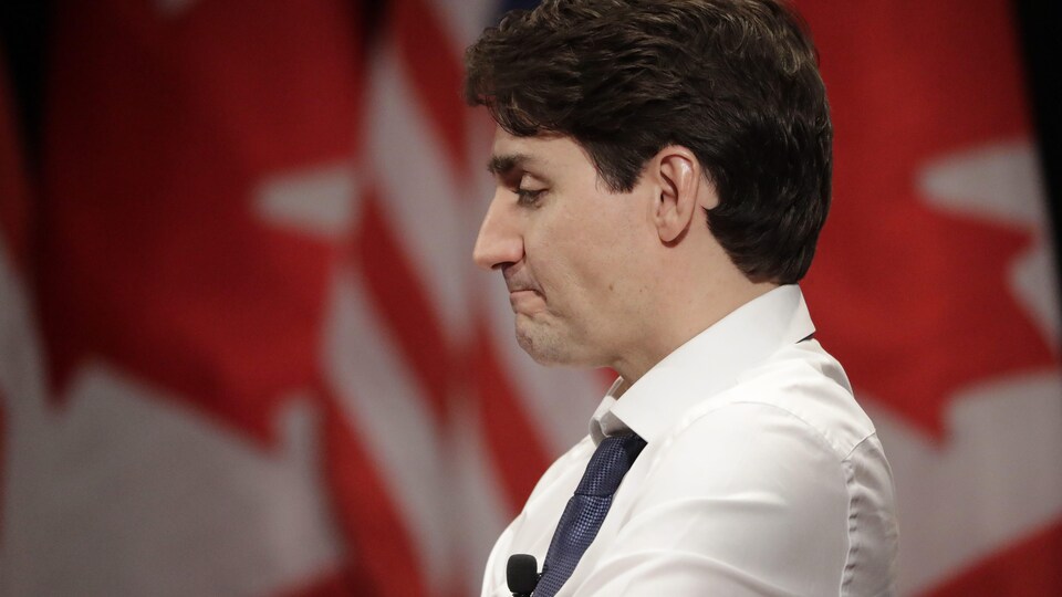 Le premier ministre Justin Trudeau apparaît de profil, vêtu d'une chemise blanche avec cravate bleue, devant des drapeaux unifoliés