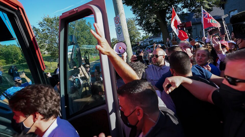 Justin Trudeau remonte dans l'autocar du Parti libéral, entouré de son équipe de sécurité. Autour se trouvent des manifestants qui brandissent des pancartes, crient ou font un doigt d'honneur.