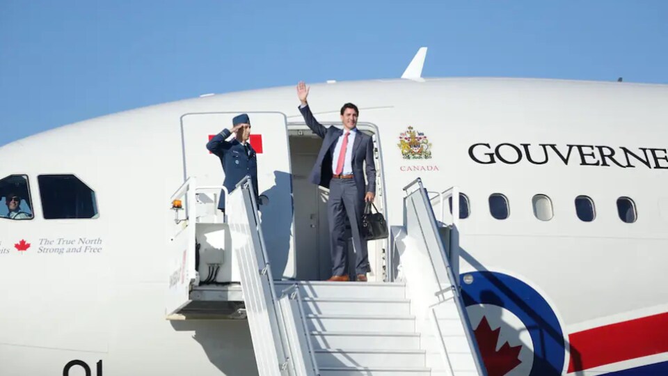 Justin Trudeau tient une valise et fait un salut de la main.
