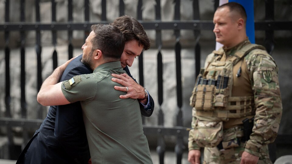Volodymyr Zelensky and Justin Trudeau hug each other in front of a soldier.