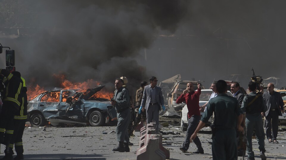 Plusieurs personnes dont des militaires et des secouristes sont sur les lieux de l'attentat, où une voiture en flamme laisse échapper beaucoup de fumée.