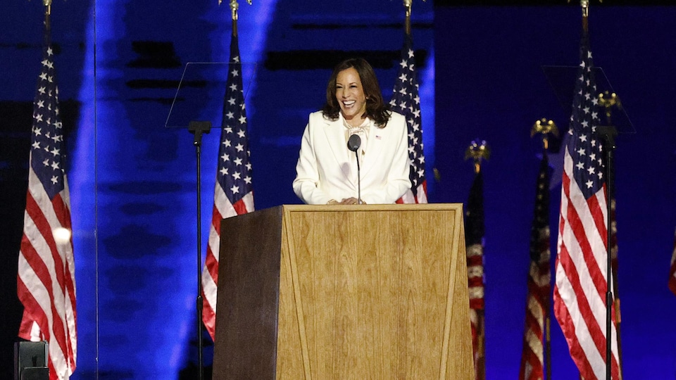 Kamala Harris est tout sourire derrière un lutrin sur une scène.