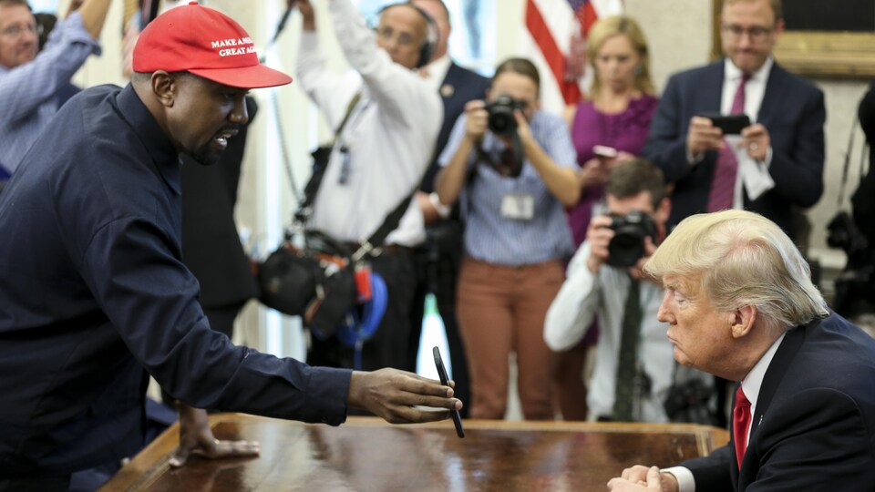 Un homme portant une casquette rouge montre son téléphone au président donald trump, assis derrière le bureau ovale, à la maison-Blanche. 